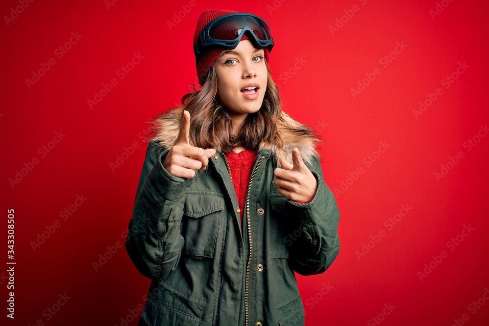 Young blonde girl wearing ski glasses and winter coat for ski weather over red background pointing fingers to camera with happy and funny face. Good energy and vibes. Young blonde girl wearing ski glasses and winter coat for ski weather over red background pointing fingers to camera with happy and funny face. Good energy and vibes.