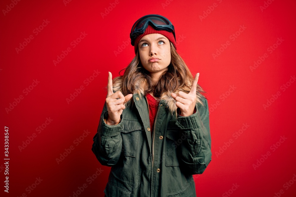 Young blonde girl wearing ski glasses and winter coat for ski weather over red background Pointing up looking sad and upset, indicating direction with fingers, unhappy and depressed. Young blonde girl wearing ski glasses and winter coat for ski weather over red background Pointing up looking sad and upset, indicating direction with fingers, unhappy and depressed.