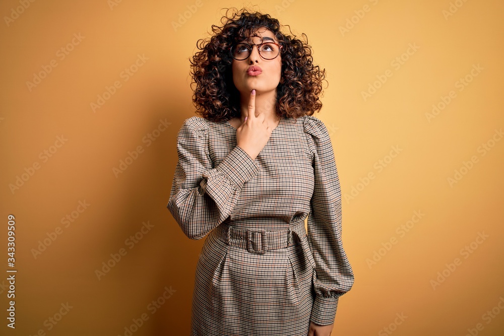 Beautiful arab business woman wearing dress and glasses standing over yellow background Thinking concentrated about doubt with finger on chin and looking up wondering