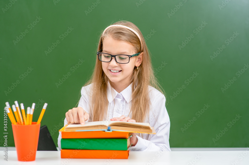 Smart smiling girl wearing eyeglasses reads book near empty green chalkboard. Empty space for text Smart smiling girl wearing eyeglasses reads book near empty green chalkboard. Empty space for text