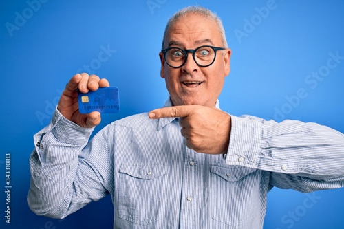 Middle age hoary man wearing glasses holding credit card as money to do payment very happy pointing with hand and finger