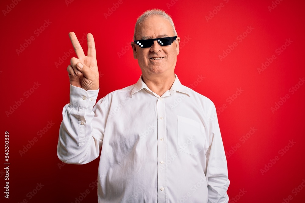 Naklejka premium Middle age hoary man wearing funny sunglasses over isolated red background smiling with happy face winking at the camera doing victory sign. Number two.