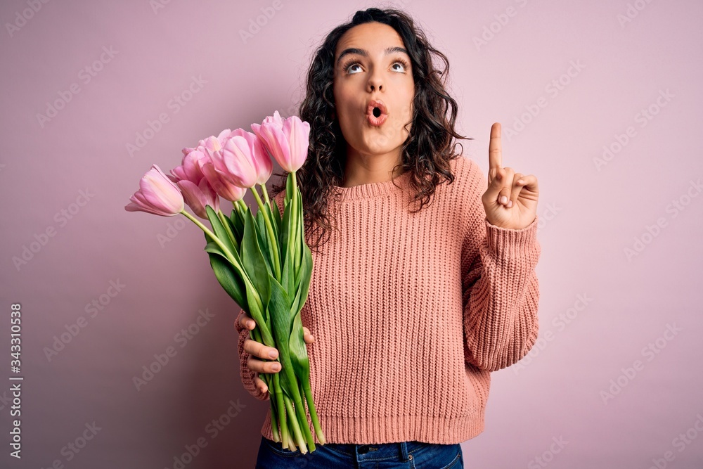 Young beautiful romantic woman with curly hair holding bouquet of pink tulips amazed and surprised looking up and pointing with fingers and raised arms. Young beautiful romantic woman with curly hair holding bouquet of pink tulips amazed and surprised looking up and pointing with fingers and raised arms.