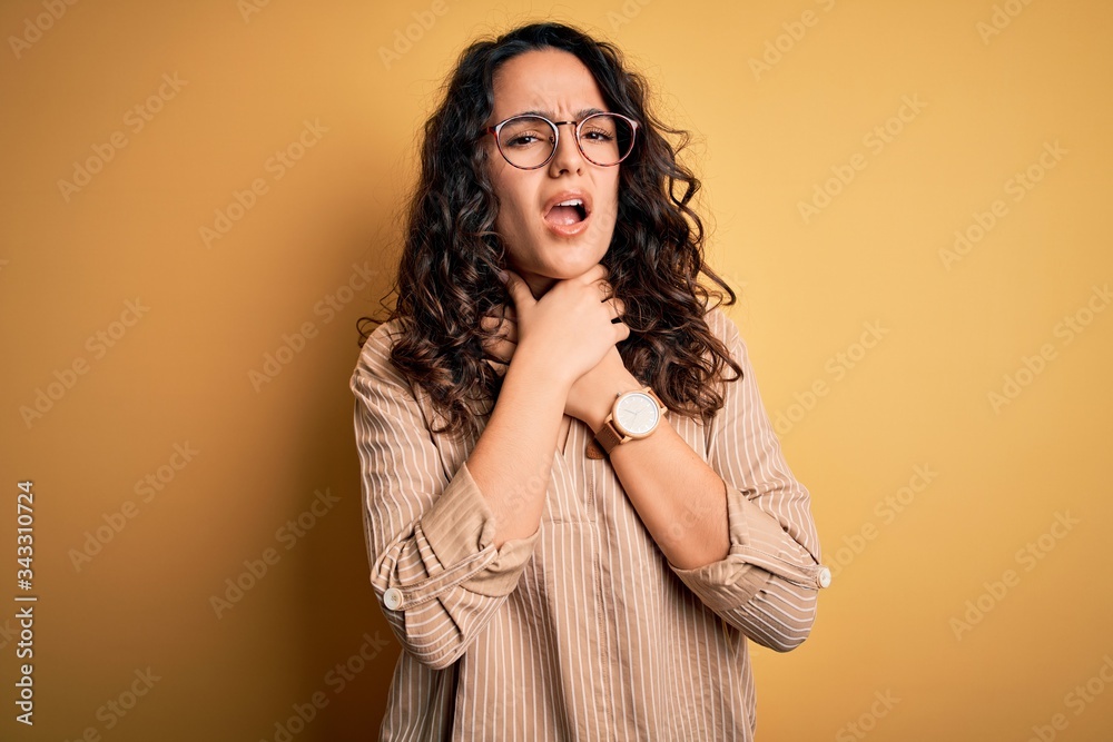 Beautiful woman with curly hair wearing striped shirt and glasses over yellow background shouting suffocate because painful strangle. Health problem. Asphyxiate and suicide concept. Beautiful woman with curly hair wearing striped shirt and glasses over yellow background shouting suffocate because painful strangle. Health problem. Asphyxiate and suicide concept.