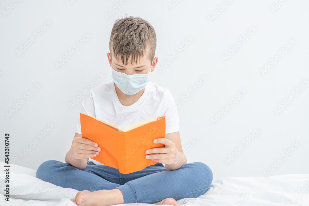 Smart boy wearing medical mask sits on a bed and reads a book. Quarantine and coronavirus epidemic concept. Empty space for text