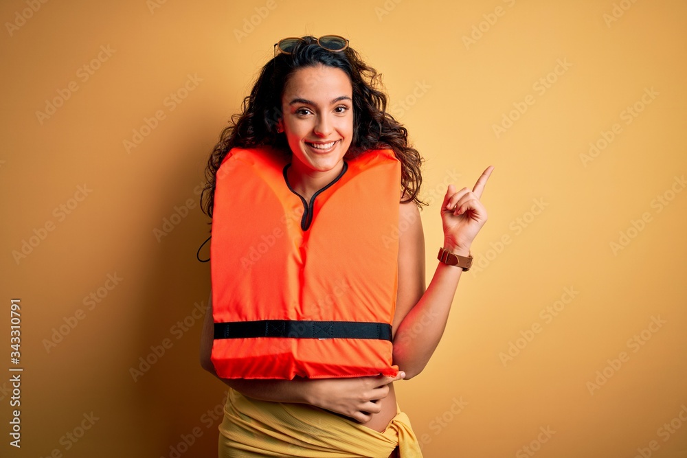 Young beautiful woman with curly hair wearing orange lifejacket over yellow background with a big smile on face, pointing with hand and finger to the side looking at the camera. Young beautiful woman with curly hair wearing orange lifejacket over yellow background with a big smile on face, pointing with hand and finger to the side looking at the camera.