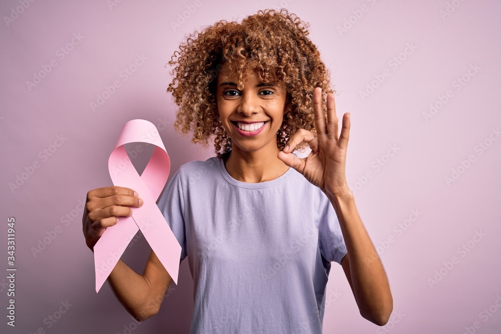 Young african american woman with curly hair holding pink cancer ribbon symbol doing ok sign with fingers, excellent symbol