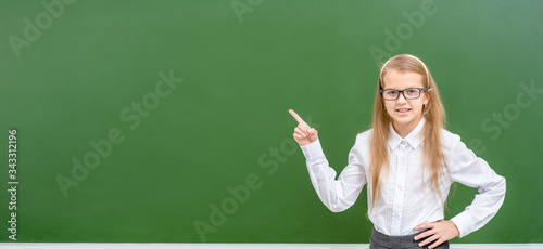 Smiling girl wearing eyeglasses points away on empty green blackboard. Empty space for text