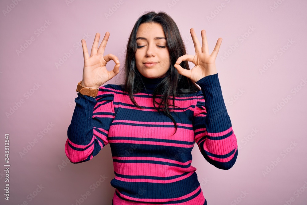 Young brunette elegant woman wearing striped shirt over pink isolated background relax and smiling with eyes closed doing meditation gesture with fingers. Yoga concept. Young brunette elegant woman wearing striped shirt over pink isolated background relax and smiling with eyes closed doing meditation gesture with fingers. Yoga concept.