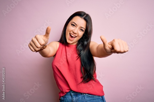 Young brunette woman wearing casual summer shirt over pink isolated background approving doing positive gesture with hand, thumbs up smiling and happy for success. Winner gesture.