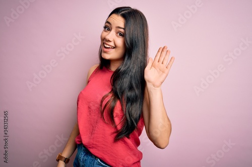 Young brunette woman wearing casual summer shirt over pink isolated background Waiving saying hello happy and smiling, friendly welcome gesture photo