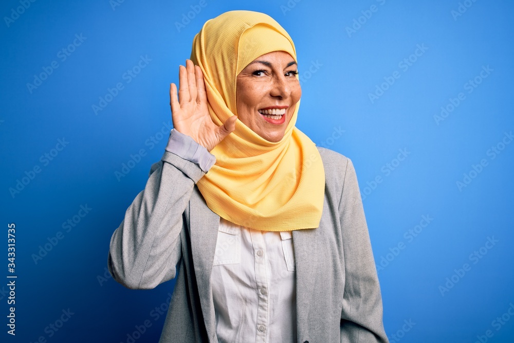 Middle age brunette business woman wearing muslim traditional hijab over blue background smiling with hand over ear listening an hearing to rumor or gossip. Deafness concept.