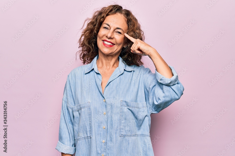 Middle age beautiful woman wearing casual denim shirt standing over pink background Smiling pointing to head with one finger, great idea or thought, good memory Middle age beautiful woman wearing casual denim shirt standing over pink background Smiling pointing to head with one finger, great idea or thought, good memory