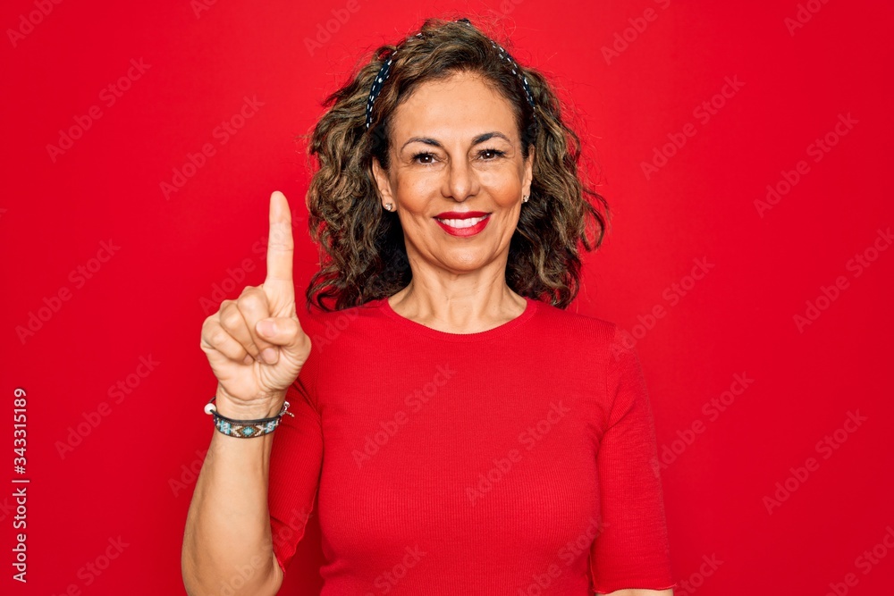 Middle age senior brunette woman wearing casual t-shirt standing over red background showing and pointing up with finger number one while smiling confident and happy. Middle age senior brunette woman wearing casual t-shirt standing over red background showing and pointing up with finger number one while smiling confident and happy.