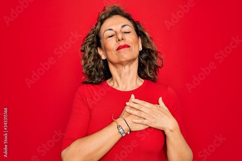Middle age senior brunette woman wearing casual t-shirt standing over red background smiling with hands on chest with closed eyes and grateful gesture on face. Health concept.