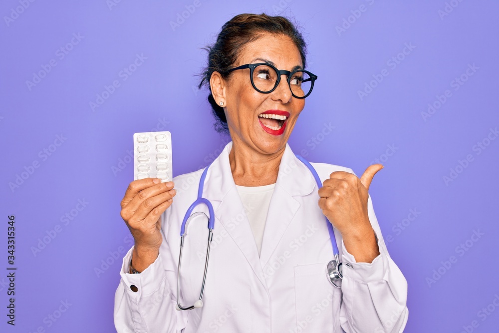 Middle age senior professional doctor woman holding pharmaceutical pills pointing and showing with thumb up to the side with happy face smiling