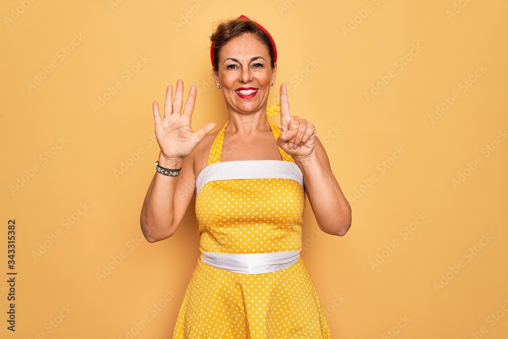 Middle age senior pin up woman wearing 50s style retro dress over yellow background showing and pointing up with fingers number six while smiling confident and happy. Middle age senior pin up woman wearing 50s style retro dress over yellow background showing and pointing up with fingers number six while smiling confident and happy.