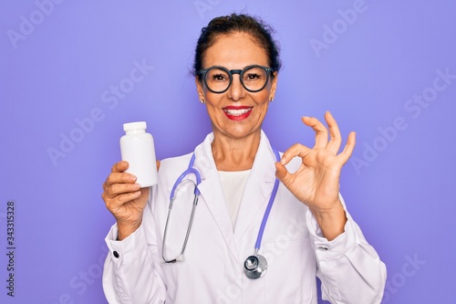Middle age senior professional doctor woman holding pharmaceutical pills doing ok sign with fingers, excellent symbol