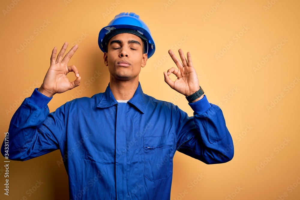 Young handsome african american worker man wearing blue uniform and security helmet relax and smiling with eyes closed doing meditation gesture with fingers. Yoga concept.