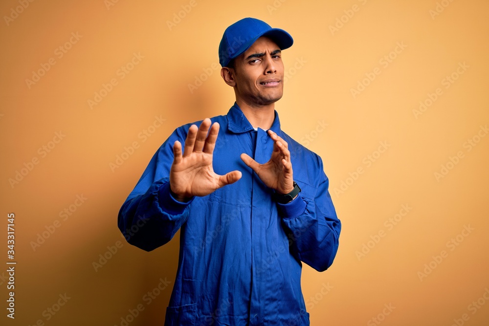 Young african american mechanic man wearing blue uniform and cap over yellow background disgusted expression, displeased and fearful doing disgust face because aversion reaction. With hands raised