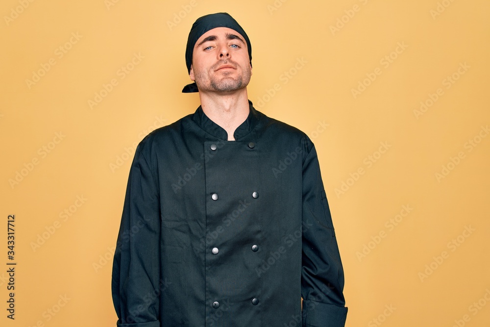 Young handsome cooker man with blue eyes wearing uniform and hat over yellow background Relaxed with serious expression on face. Simple and natural looking at the camera. Young handsome cooker man with blue eyes wearing uniform and hat over yellow background Relaxed with serious expression on face. Simple and natural looking at the camera.