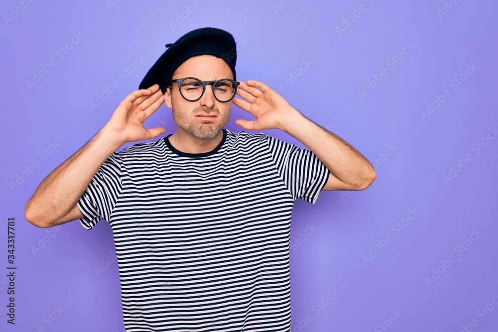 Naklejka premium Handsome man with blue eyes wearing striped t-shirt and french beret over purple background Trying to hear both hands on ear gesture, curious for gossip. Hearing problem, deaf