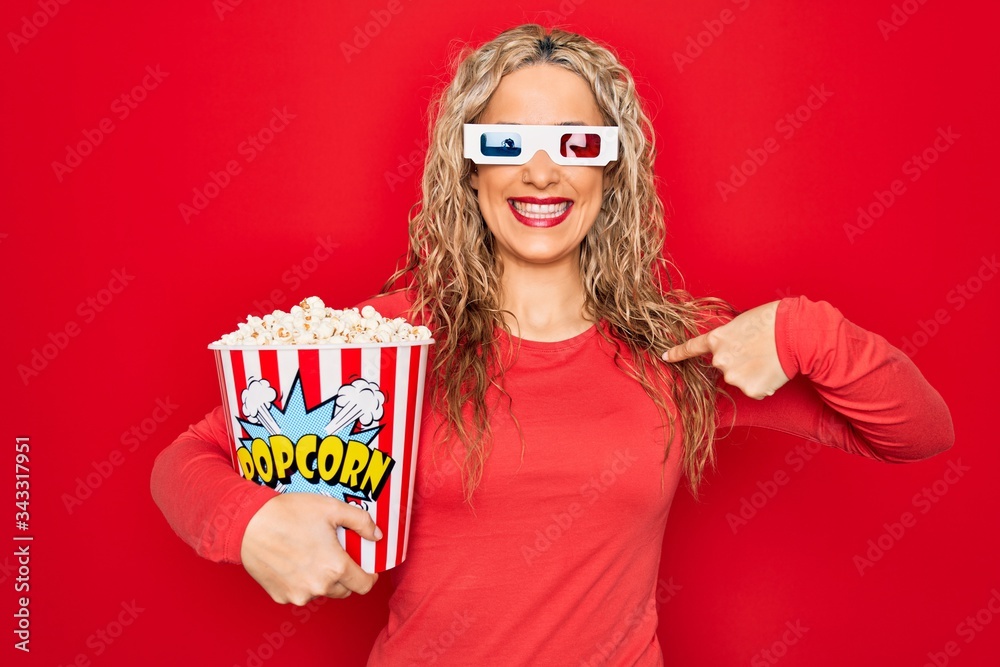 Obraz premium Young beautiful blonde woman watching movie using 3d glasses eating popcorn pointing finger to one self smiling happy and proud