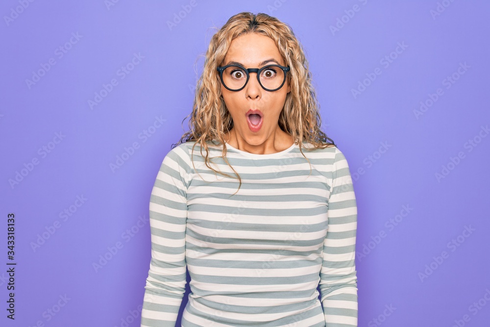 Beautiful blonde woman wearing casual striped t-shirt and glasses over purple background afraid and shocked with surprise and amazed expression, fear and excited face. Beautiful blonde woman wearing casual striped t-shirt and glasses over purple background afraid and shocked with surprise and amazed expression, fear and excited face.