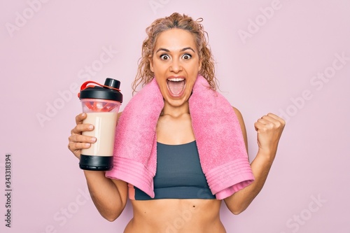 Beautiful blonde sporty woman doing sport wearing towel holding shaker with protein beverage screaming proud, celebrating victory and success very excited with raised arms photo