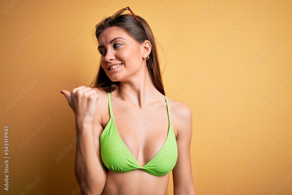 Young beautiful woman with blue eyes on vacation wearing bikini and sunglasses smiling with happy face looking and pointing to the side with thumb up. Young beautiful woman with blue eyes on vacation wearing bikini and sunglasses smiling with happy face looking and pointing to the side with thumb up.