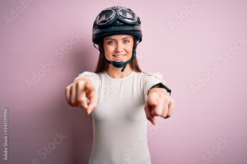 Young beautiful motorcyclist woman with blue eyes wearing moto helmet over pink background pointing to you and the camera with fingers, smiling positive and cheerful