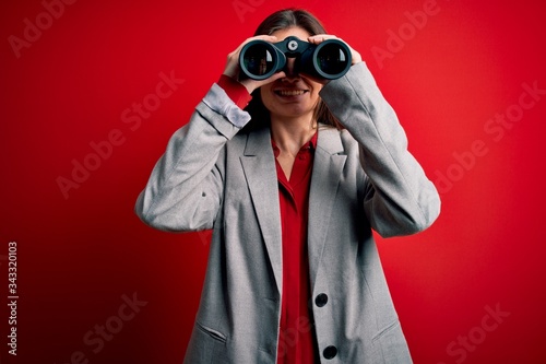 Young beautiful woman with blue eyes using binoculars over isolated red background with a happy face standing and smiling with a confident smile showing teeth