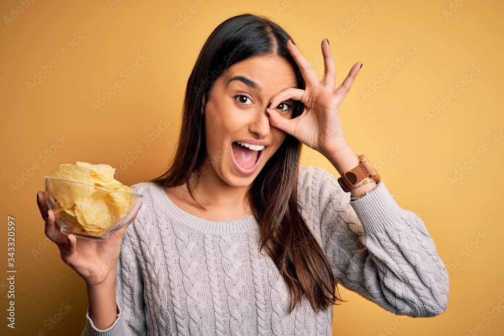 Young beautiful brunette woman holding bowl with snack potato chips over yellow background with happy face smiling doing ok sign with hand on eye looking through fingers Young beautiful brunette woman holding bowl with snack potato chips over yellow background with happy face smiling doing ok sign with hand on eye looking through fingers