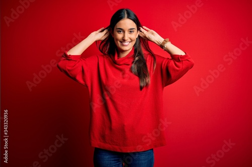 Young beautiful brunette woman wearing casual sweater over isolated red background Smiling pulling ears with fingers, funny gesture. Audition problem © Krakenimages.com