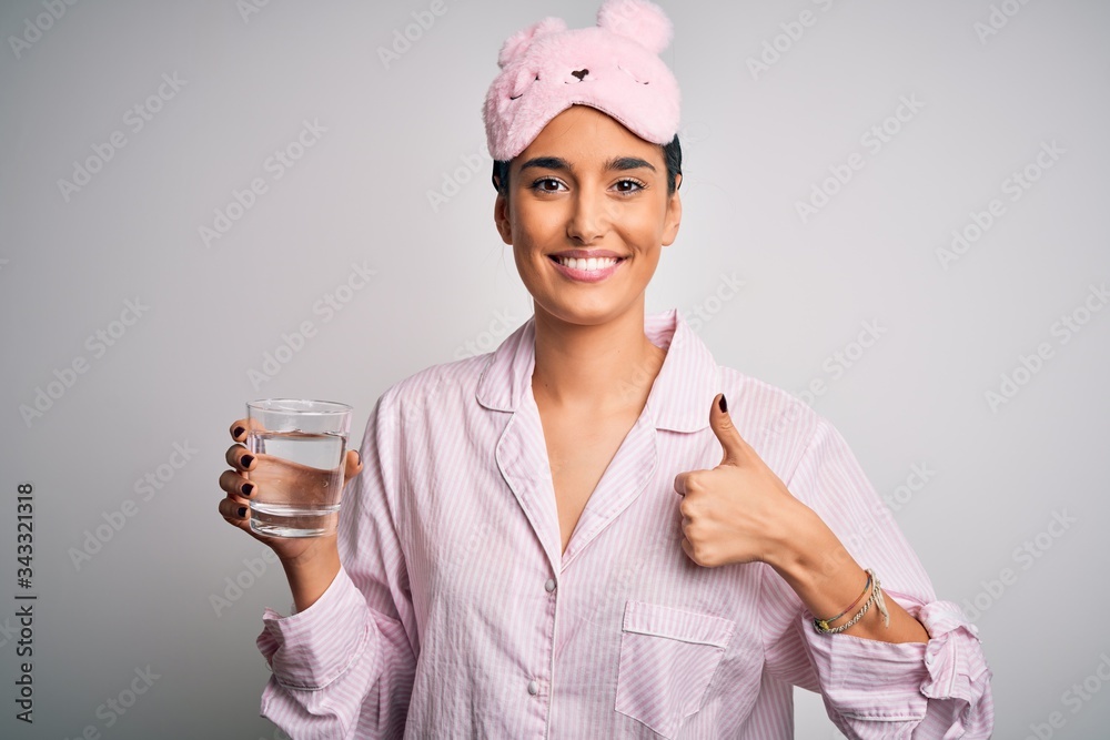 Young beautiful woman wearing pajama and sleep mask drinking glass of water happy with big smile doing ok sign, thumb up with fingers, excellent sign Young beautiful woman wearing pajama and sleep mask drinking glass of water happy with big smile doing ok sign, thumb up with fingers, excellent sign
