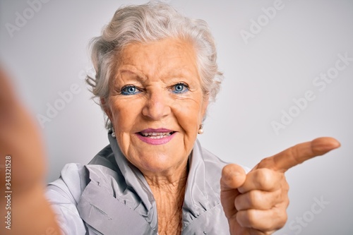 Senior beautiful grey-haired woman making selfie by camera over isolated white background very happy pointing with hand and finger to the side