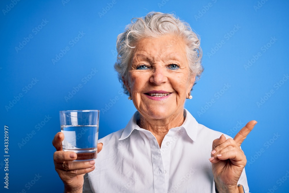 Senior beautiful woman drinking glass of water standing over isolated blue background very happy pointing with hand and finger to the side Senior beautiful woman drinking glass of water standing over isolated blue background very happy pointing with hand and finger to the side