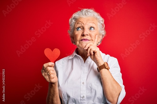 Senior beautiful woman holding paper heart standing over isolated red background serious face thinking about question, very confused idea