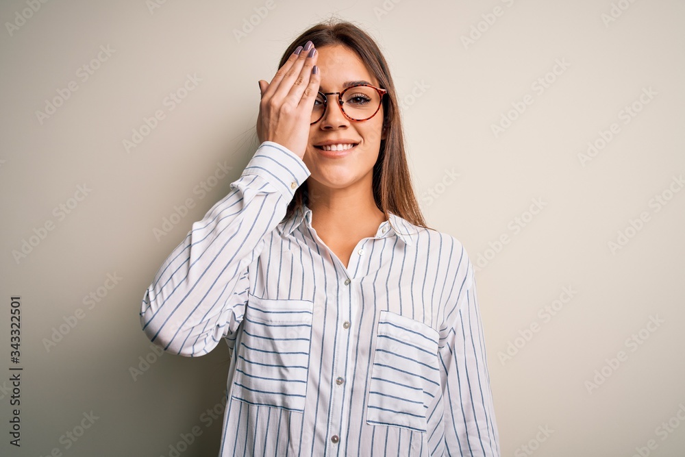 Young beautiful brunette woman wearing casual shirt and glasses over white background covering one eye with hand, confident smile on face and surprise emotion. Young beautiful brunette woman wearing casual shirt and glasses over white background covering one eye with hand, confident smile on face and surprise emotion.
