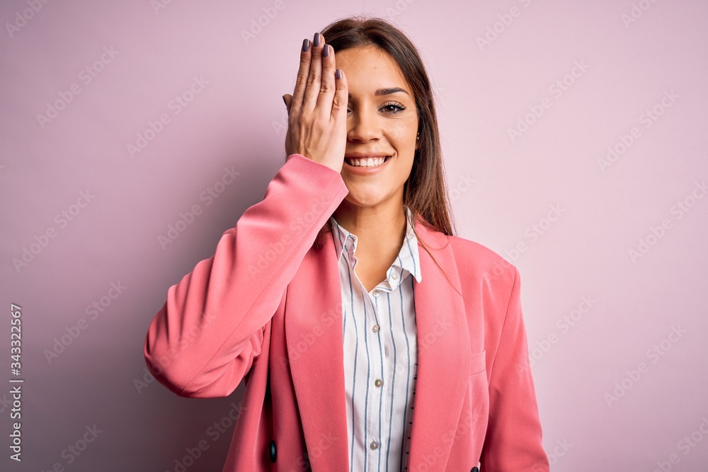 Young beautiful brunette woman wearing jacket standing over isolated pink background covering one eye with hand, confident smile on face and surprise emotion. Young beautiful brunette woman wearing jacket standing over isolated pink background covering one eye with hand, confident smile on face and surprise emotion.