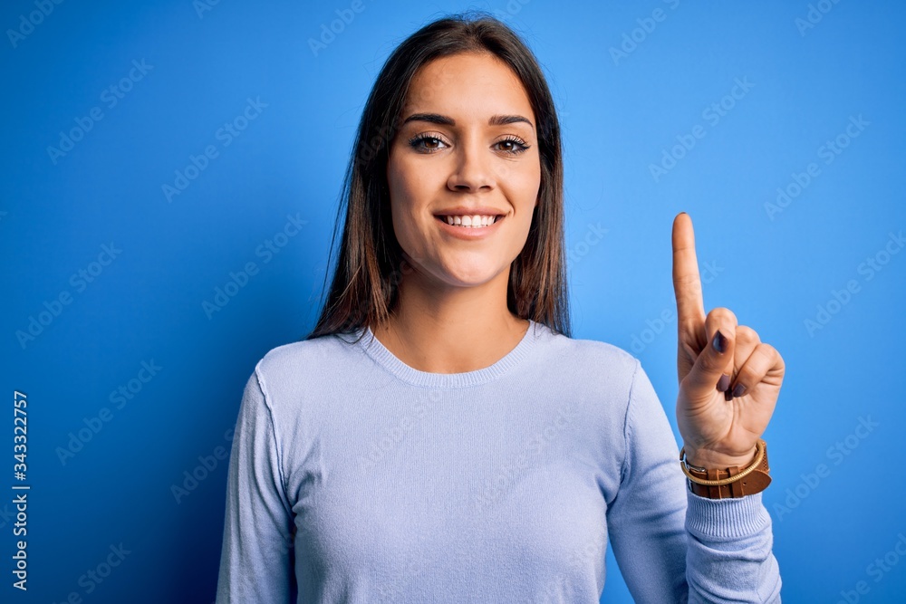 Young beautiful brunette woman wearing casual sweater standing over blue background pointing finger up with successful idea. Exited and happy. Number one.