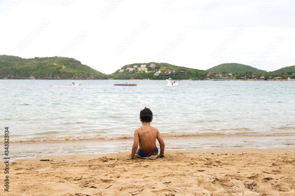 A lonely brazilian boy sitting on the sand on a beach of Brazil. View of his backs. Ferradura Beach, Buzios.