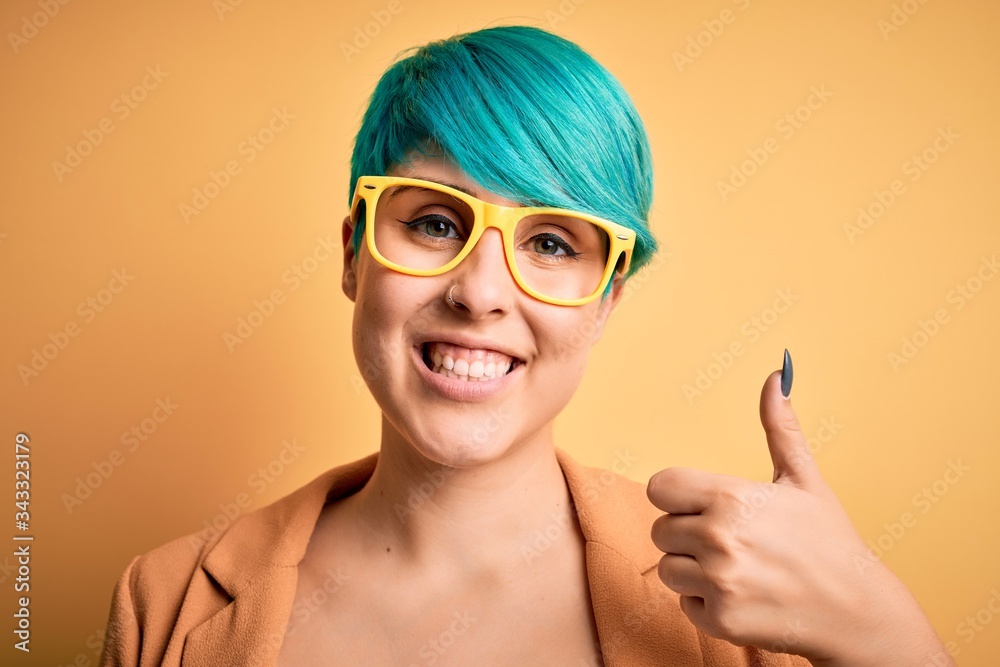 Fototapeta premium Young beautiful woman with blue fashion hair wearing casual glasses over yellow background happy with big smile doing ok sign, thumb up with fingers, excellent sign