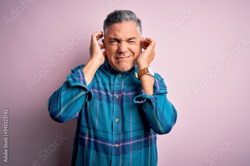 Middle age handsome grey-haired man wearing casual shirt over isolated pink background covering ears with fingers with annoyed expression for the noise of loud music. Deaf concept. © Krakenimages.com