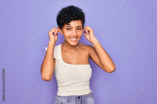 Young beautiful african american afro woman wearing casual t-shirt over purple background Smiling pulling ears with fingers  funny gesture. Audition problem