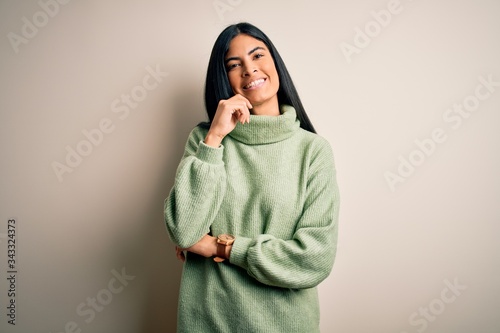 Young beautiful hispanic woman wearing green winter sweater over isolated background looking confident at the camera smiling with crossed arms and hand raised on chin. Thinking positive. © Krakenimages.com