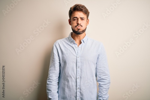 Young handsome man with beard wearing striped shirt standing over white background puffing cheeks with funny face. Mouth inflated with air, crazy expression.