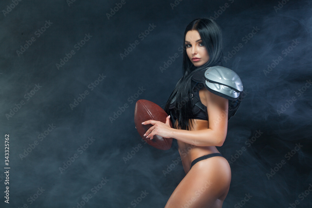 Fototapeta American football. Young sporty brunette wearing sexy uniform of rugby football player posing with ball in smoke isolated on black background