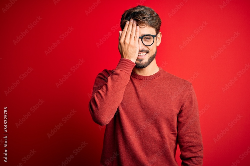 Young handsome man with beard wearing glasses and sweater standing over red background covering one eye with hand, confident smile on face and surprise emotion. Young handsome man with beard wearing glasses and sweater standing over red background covering one eye with hand, confident smile on face and surprise emotion.