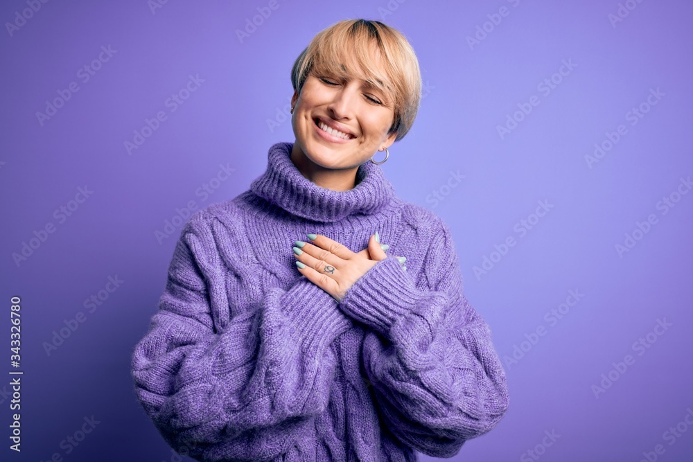 Young blonde woman with short hair wearing winter turtleneck sweater over purple background smiling with hands on chest with closed eyes and grateful gesture on face. Health concept. Young blonde woman with short hair wearing winter turtleneck sweater over purple background smiling with hands on chest with closed eyes and grateful gesture on face. Health concept.